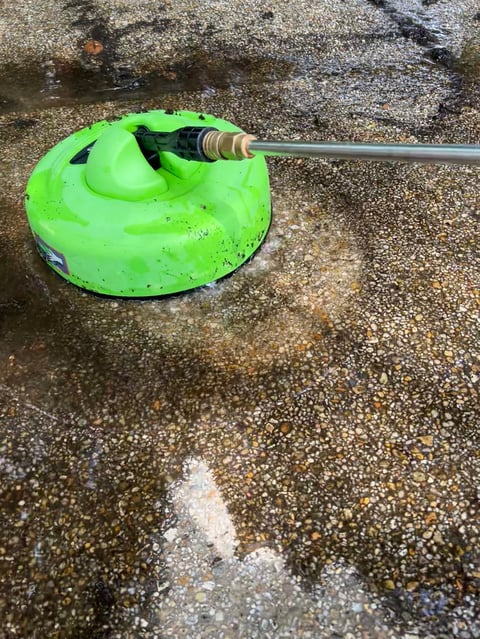The image shows a power washer cleaning a concrete surface, with water spraying onto the ground through a green surface cleaner attachment.