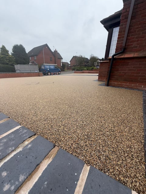 Residential driveway with gravel surface, brick houses, parked car, and overcast sky in suburban neighborhood