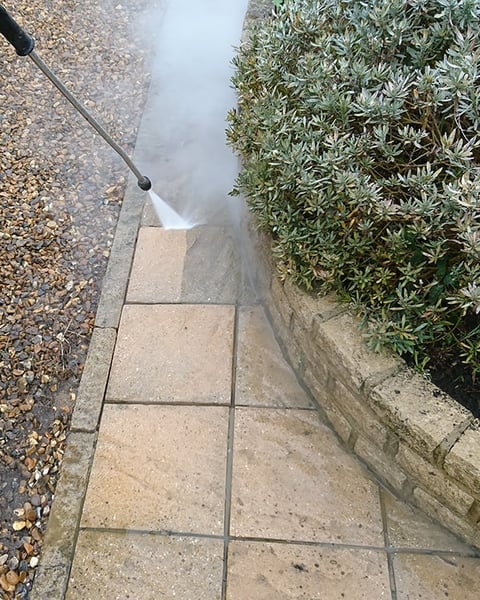 Pressure washing a paved pathway between gravel and green hedging with water spray visible