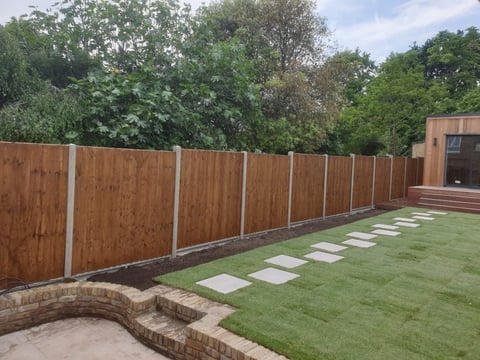 Residential backyard with wooden fence, manicured green lawn, stepping stone pathway, brick retaining wall, and trees in background