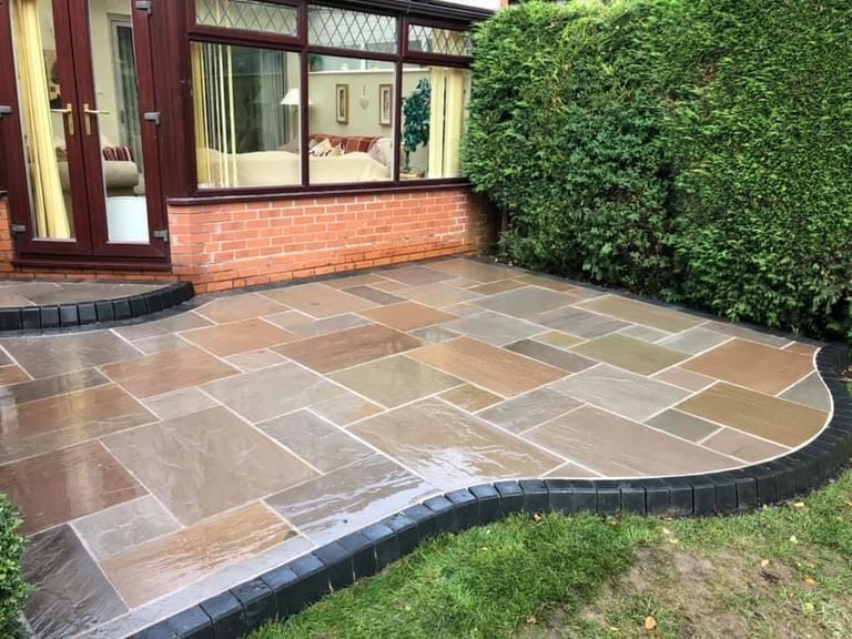 Curved patio with stone tiles bordered by black edging, adjacent to brick house with ivy-covered wall