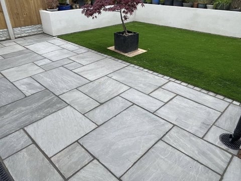 Modern patio with gray stone pavers in herringbone pattern, bordered by white walls, green lawn, and a potted tree in the background