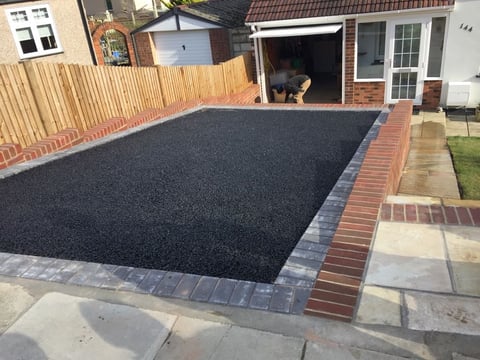 Newly constructed driveway with dark asphalt surface and red brick border in residential backyard, with house and garage visible