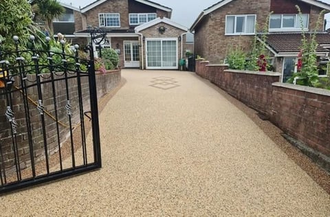 Residential driveway with gravel surface, black metal gate on left, brick houses with gardens on sides